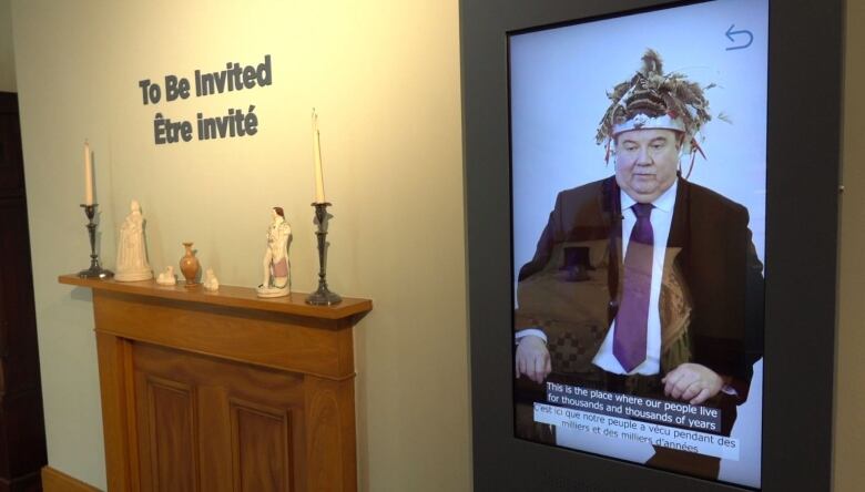 A man wearing a Mohawk headdress is shown on a video screen hanging from a museum wall.