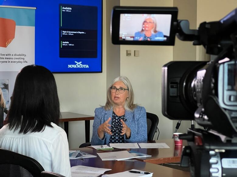 Two women sit facing each other in a boardroom. One woman is gesturing and explaining something to the other. A camera is in the foreground, videotaping the interview. 