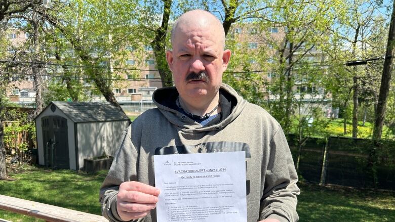 A bald man with moustache wearing a hoodie holds up a piece of paper in his backyard.