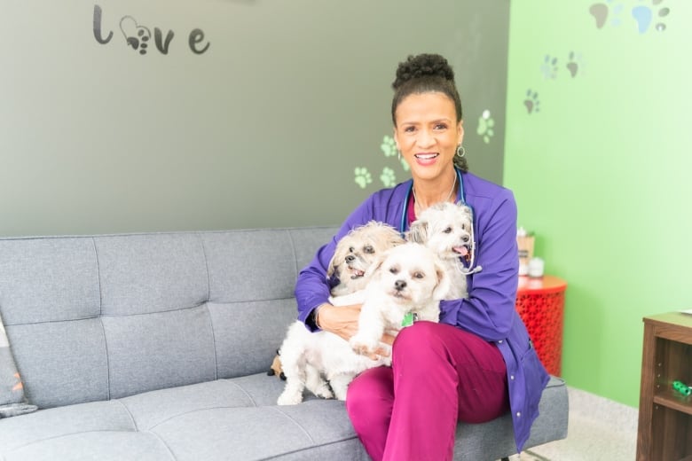 A black woman with her hair pulled back wearing a purple sweater and red medical scrubs sits on a couch holding three small flufly white dogs.