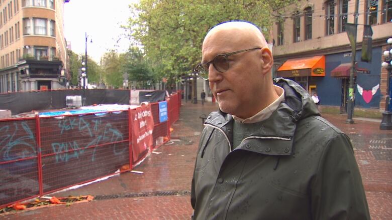 Man with short grey hair wearing glasses stands in rainy downtown street in front of construction barrier.