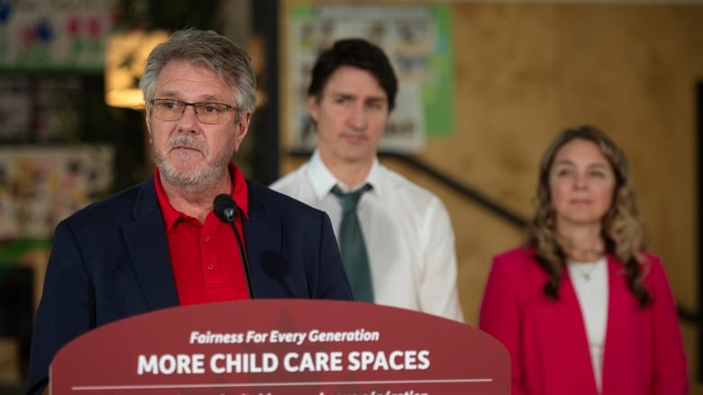 A man, with another man and a woman behind him, speaks at a podium.