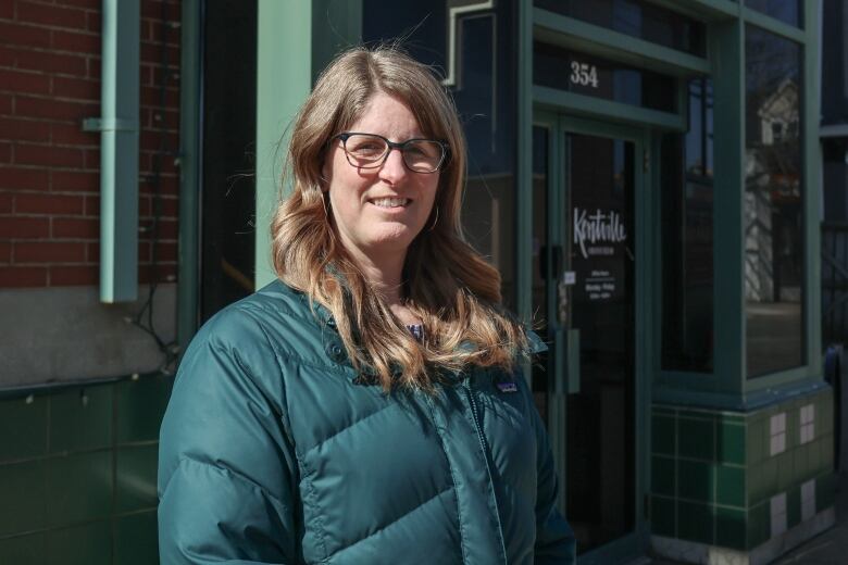 A blonde woman with long hair and glasses stands outside the door of a building. She is wearing a puffy dark green jacket. The building door is made of glass and has the word 