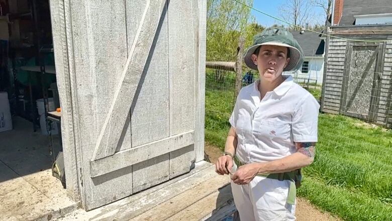 A woman, wearing a white jumpsuit and a hat, stands in front of a barn. 