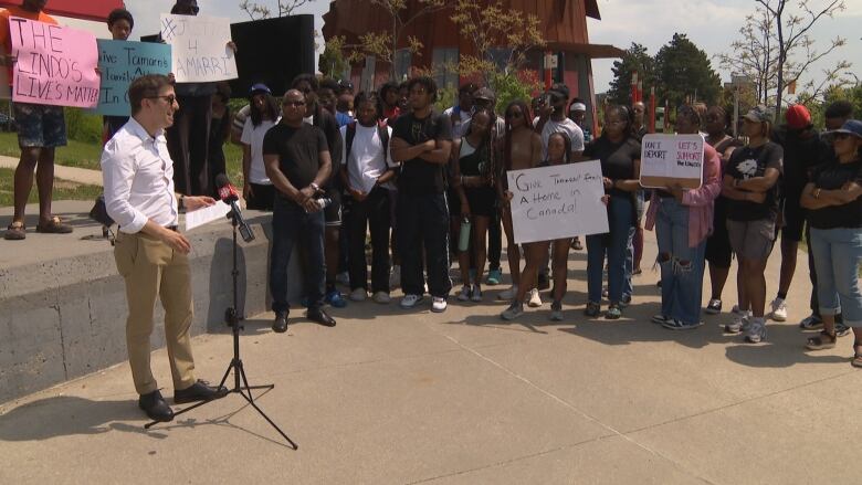 A white man speaks into a microphone at a rally attended by dozens of Black people of all ages. People in the crowd are holding signs supporting the Lindo family's right to stay in Canada