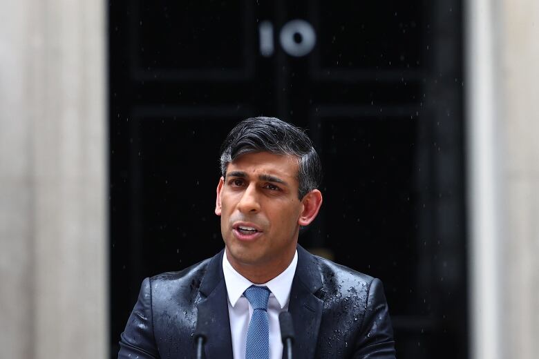 A man in a suit and tie whose clothing is wet speaks outside.