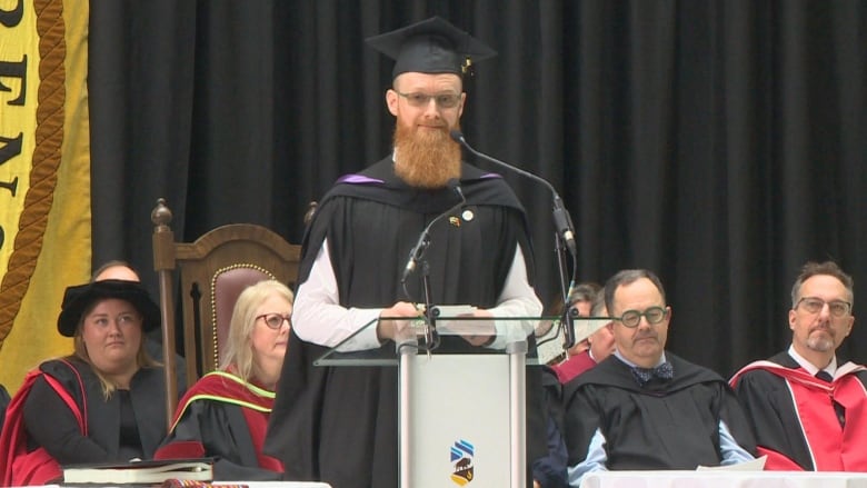 A man is pictured giving a speech at a podium, wearing a graduation gown.