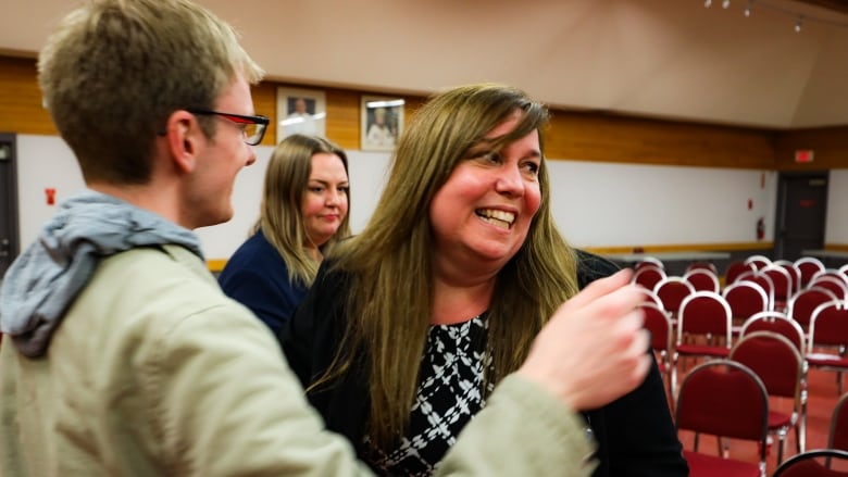 A woman in a black blazer smiles and looks over to the side, after receiving a hug.