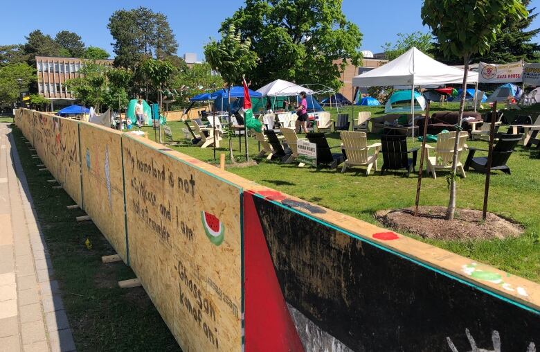 A barricade made of plywood between a sidewalk and a greenspace as part of a protest. There are tents and chairs in the greenspace.