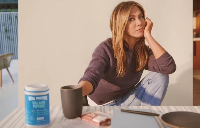 A woman sits behind a table looking at the camera. On the table is a mug, protein bar and blue tub of collagen powder.