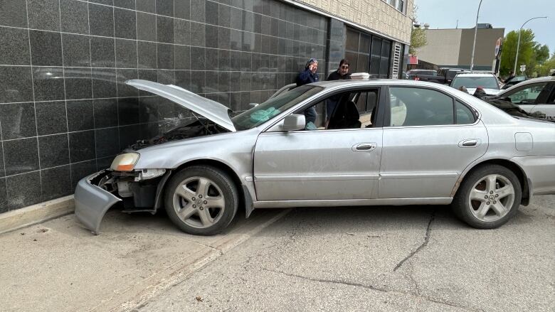 A silver vehicle can be seen with its front bumper laying on the ground. The car's hood is up and scratches can be seen on the building's exterior. 