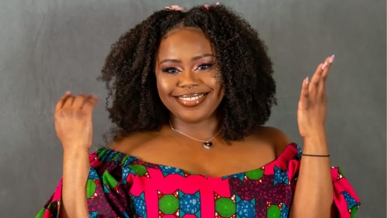 A woman with shoulder-length curling hair wears a heart necklace and a patterned off-the-shoulders top.