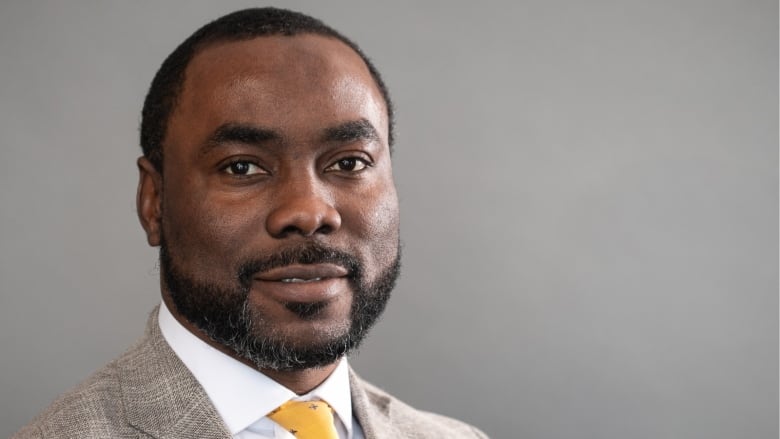 Man wearing a grey suit with a white shirt and yellow tie stands in front of a grey background.