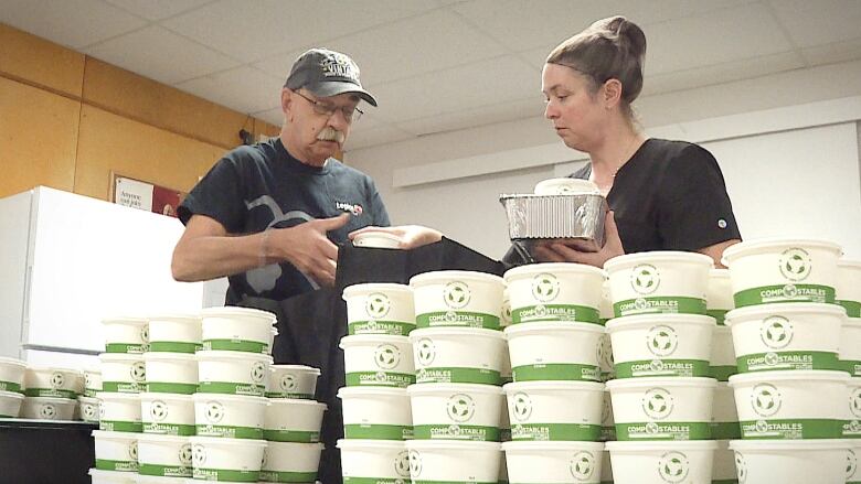 A man hands a cardboard soup carton to a woman, both of whom are behind a long stack of soup containers.