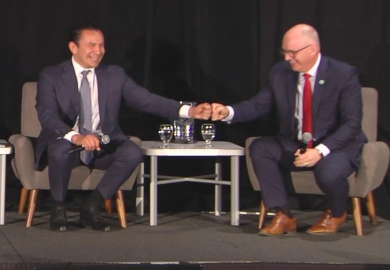 Two men wearing suits are sitting on a stage and fist bumping each other, while laughing. A third man wearing a suit is seated to the left of them.