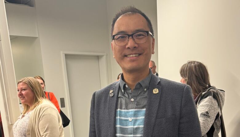 Vertical photo of a middle-aged Asian-Canadian man, wearing a blazer and jeans. Indoor photo. Several people in the background. 