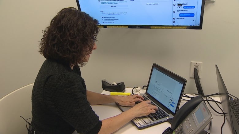 A woman with shoulder length curly black hair wearing a black top sits at a table typing on a laptop.