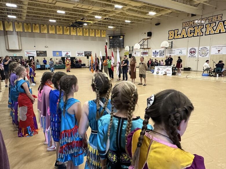 A line of young girls in colourful clothing with long braids down their backs stand in a gymnasium.