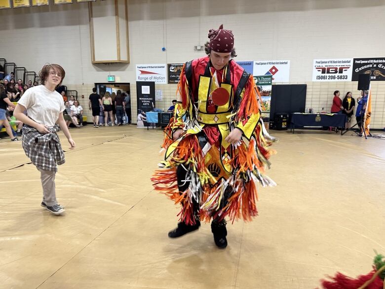 A person wearing red, yellow and orange traditional Indigenous dress dances in a gymnasium. 