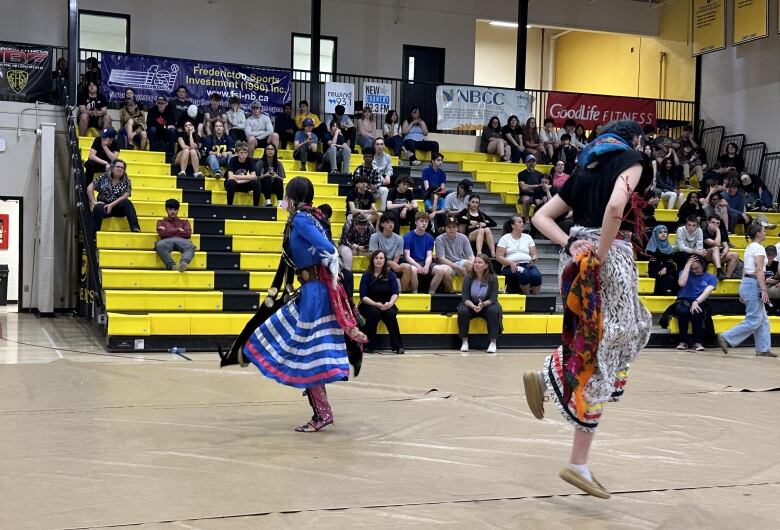 Two people wearing colourful ribbon skirts dance in a gymnasium as students watch from the bleachers.