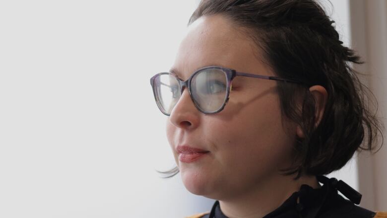 A woman with a bob haircut wearing glasses looks to the left. She is standing in front of a window.