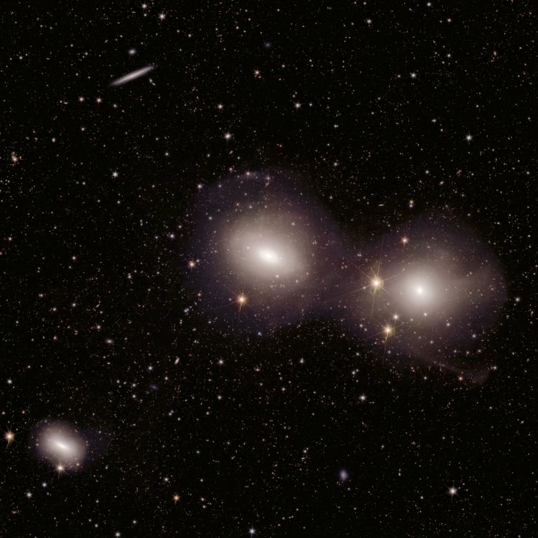 Three bright galaxies are seen in the foreground with thousands of stars and other smaller galaxies.