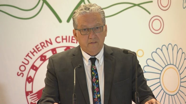 A man with wavy white hair and wearing a suit stands and talks at a podium.