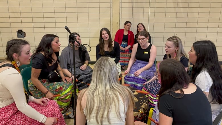 A group of teenage girls sit around a big drum, holding up sticks and singing.