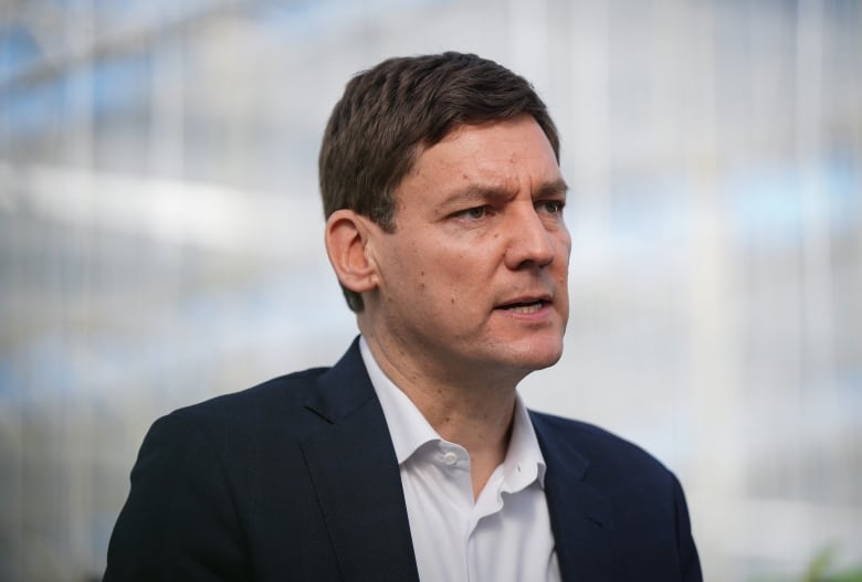 A portrait of B.C. Premier David Eby, a tall man wearing a suit and an open-collared white shirt, speaking during an announcement.