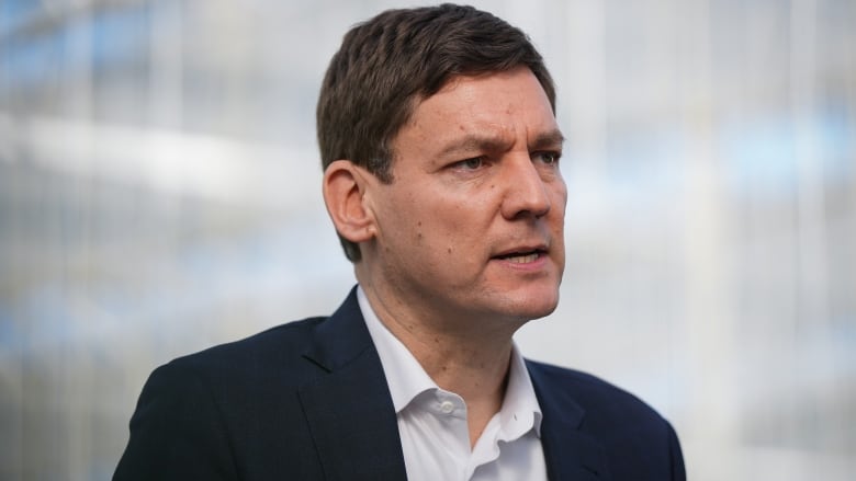 A tall man wearing a suit and an open-collared white shirt speaks during an announcement.