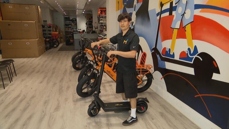 A man looks at the camera while holding an electric scooter. Other electric scooters are seen behind him.