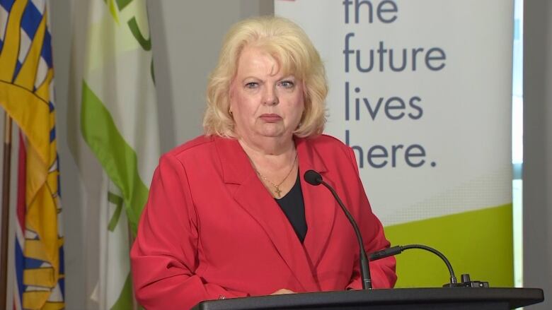 A woman with blond hair and a bright red blazer stands at a microphone.