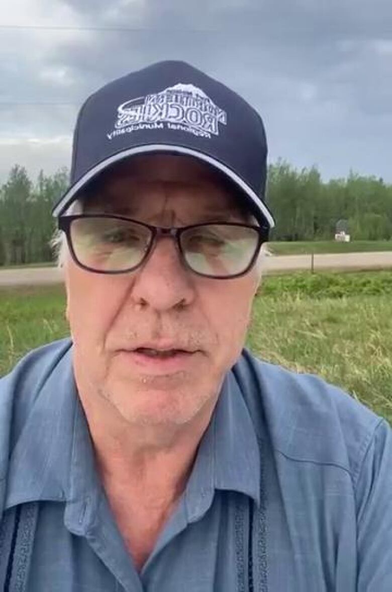 A white man wearing a blue shirt and hat stands in a field during a cloudy day.