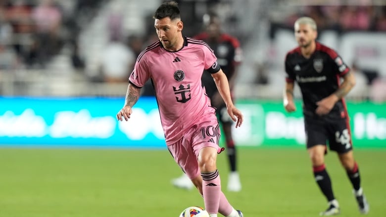 A soccer player wearing pink dribbles the ball, while others wearing black are pictured behind him.