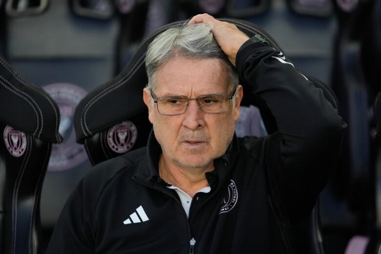 A Hispanic man scratches his head while sitting in a soccer dugout.