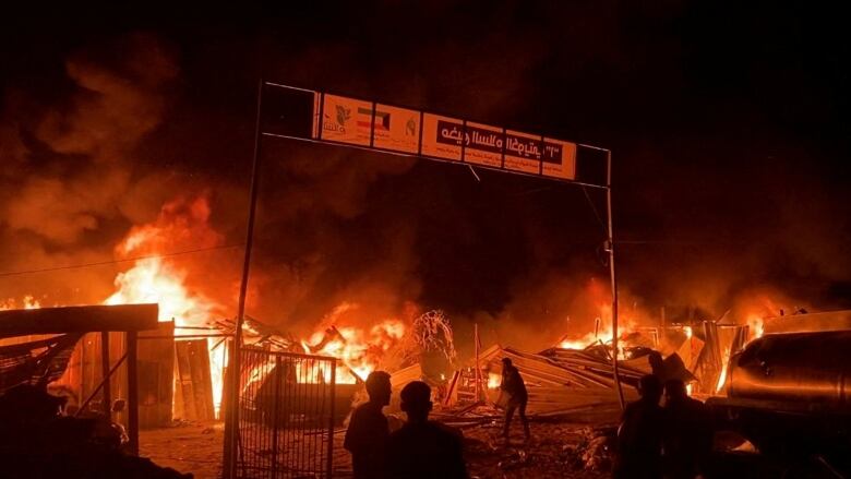 People gather around as a fire engulf  debris and destroyed buildings.