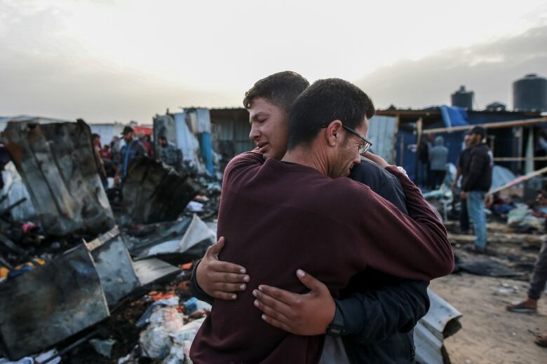 Two cleanshaven men, one wearing glasses, embrace in an outdoor photo whose background includes large pieces of debris.