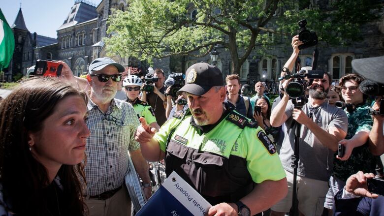 A special constable points his finger at a young woman, and they're surrounded by other people with cameras and cellphones recording the interaction.