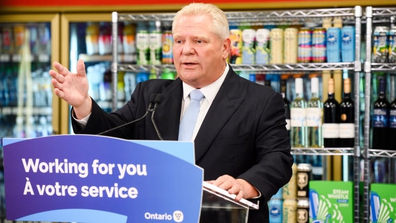 Premier Doug Ford stands at a podium with a sign saying 'Working for you,' with cans of beer and bottles of wine on shelves in the background.  