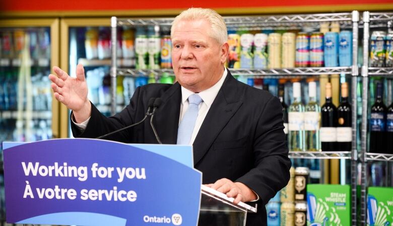 Premier Doug Ford stands at a podium with a sign saying 'Working for you,' with cans of beer and bottles of wine on shelves in the background.  