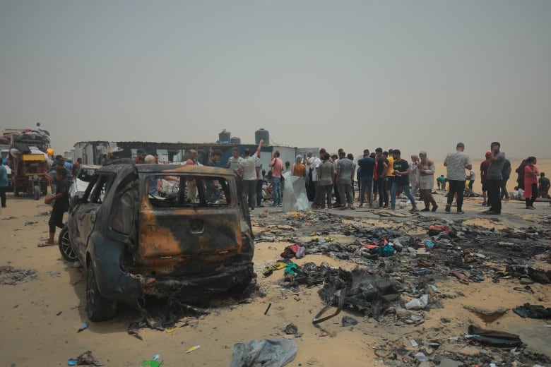 A burned car and scorched belongings are seen in what was once a camp for displaced Palestinians. A group of survivors is standing in the background.