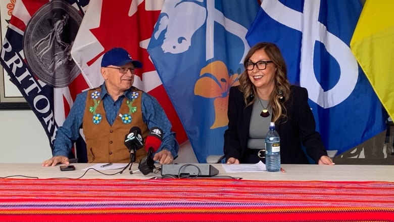 A man and a woman sit a table and speak into microphones at a media event.