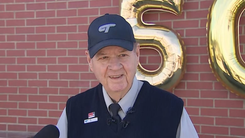 An elderly man wearing a baseball cap stands in front of a brick wall, smiling.