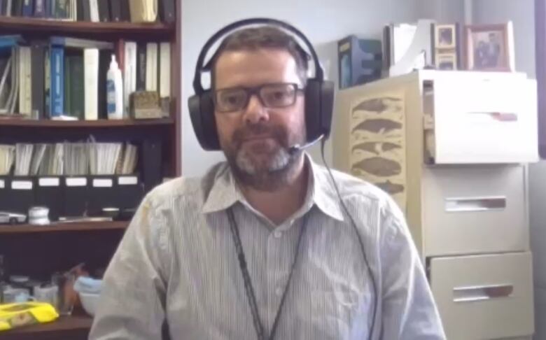 A man wearing a button-up shirt with a lanyard around his neck wears glasses. He is sitting near a filing cabinet and a bookshelf.