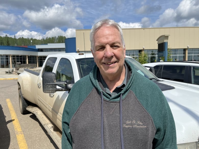 A white man wearing a green-tinged grey hoodie smiles while in a parking lot.