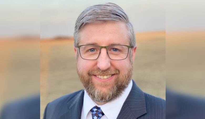 A man in glasses and a beard smiles at the camera. He is seen from the shoulders up and is wearing a suit