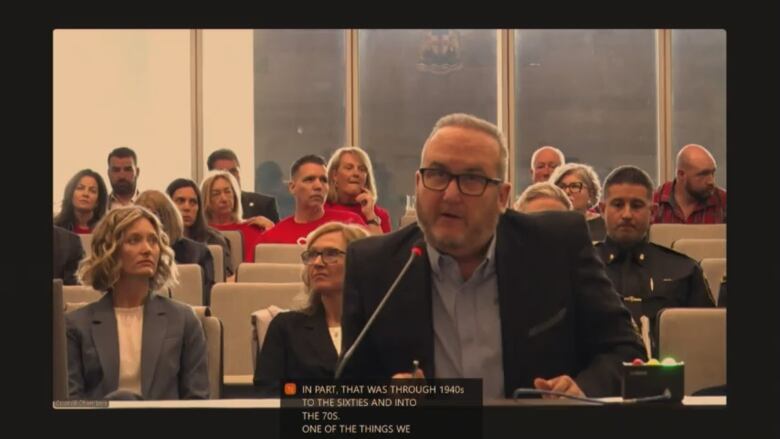 A man in a business suit wearing glasses speaks in city council chambers.