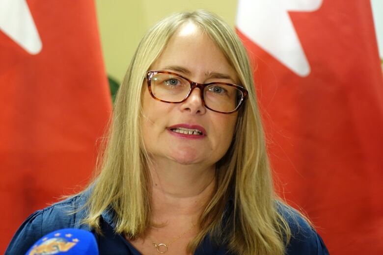 A woman with glasses and long blonde hair stands in front of two Canadian flags.