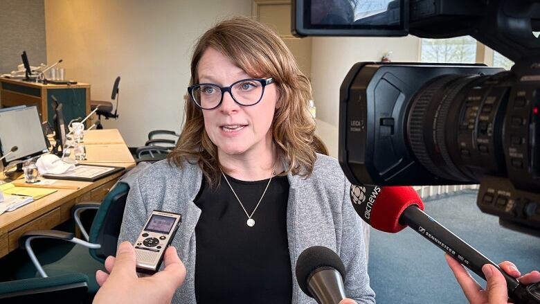 A woman with long light brown hair wearing glasses and a black top and grey jacket speaks to reporters surrounded by microphones and TV camera lenses.