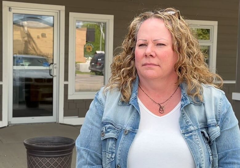 A long haired woman wearing a T-shirt, jean jacket, and necklace stands outside a building looking somber.